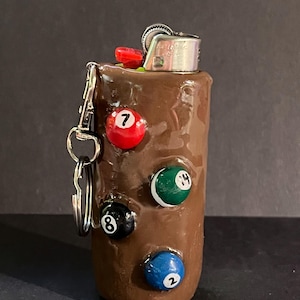 May include: A brown cylindrical lighter case featuring four embedded miniature billiard balls numbered 7, 14, 8, and 2. The case includes a silver metal keychain attachment on the side and a standard silver metal lighter mechanism at the top.