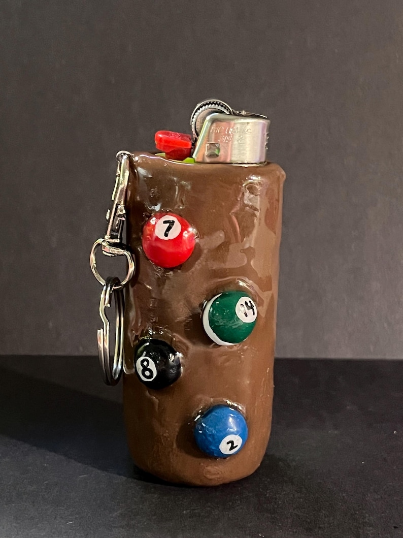May include: A brown cylindrical lighter case featuring four embedded miniature billiard balls numbered 7, 14, 8, and 2. The case includes a silver metal keychain attachment on the side and a standard silver metal lighter mechanism at the top.