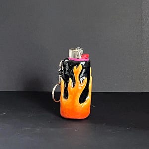 May include: A black lighter case featuring a raised, sculpted orange and yellow flame design covering the exterior. The lighter includes a metallic top mechanism with a small pink accent and a silver metal keyring attached to the side.