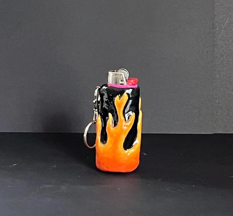 May include: A black lighter case featuring a raised, sculpted orange and yellow flame design covering the exterior. The lighter includes a metallic top mechanism with a small pink accent and a silver metal keyring attached to the side.