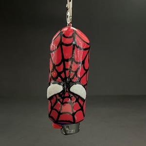 May include: A cylindrical lighter case featuring a glossy red surface with a hand-painted black spiderweb pattern and two white, almond-shaped eye accents. The case is attached to a silver-toned metal keychain ring and lobster clasp hardware.