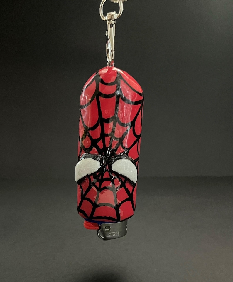 May include: A cylindrical lighter case featuring a glossy red surface with a hand-painted black spiderweb pattern and two white, almond-shaped eye accents. The case is attached to a silver-toned metal keychain ring and lobster clasp hardware.