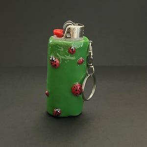 May include: A green lighter case with a glossy finish, decorated with several small red ladybugs featuring black spots. The lighter has a metallic top with a red ignition button and includes a silver metal keychain attachment on the side.