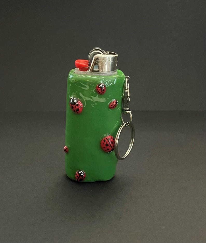 May include: A green lighter case with a glossy finish, decorated with several small red ladybugs featuring black spots. The lighter has a metallic top with a red ignition button and includes a silver metal keychain attachment on the side.
