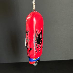 May include: A red lighter case featuring a black spider graphic and web patterns, attached to a silver metal keychain ring. The lighter inside has a blue top with the yellow Bic logo visible. The object is suspended vertically against a dark background.