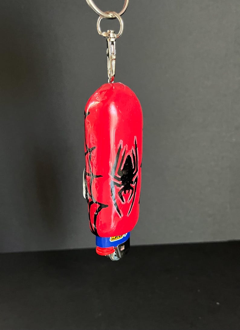 May include: A red lighter case featuring a black spider graphic and web patterns, attached to a silver metal keychain ring. The lighter inside has a blue top with the yellow Bic logo visible. The object is suspended vertically against a dark background.