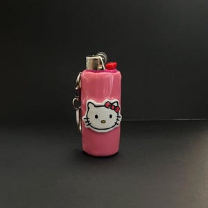 May include: A pink lighter case featuring a white Hello Kitty character face with a red bow. The cylindrical case has a metallic silver top mechanism with a red ignition button and includes a small silver metal keychain ring attached to the side.