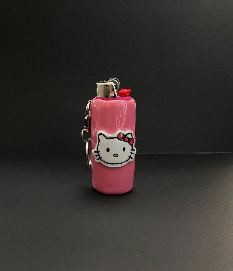 May include: A pink lighter case featuring a white Hello Kitty character face with a red bow. The cylindrical case has a metallic silver top mechanism with a red ignition button and includes a small silver metal keychain ring attached to the side.