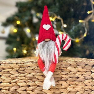 May include: A red and white gnome figurine with a candy cane in its hand. The gnome is wearing a red hat with a white heart on it and has a white beard. The gnome is sitting on a woven basket.