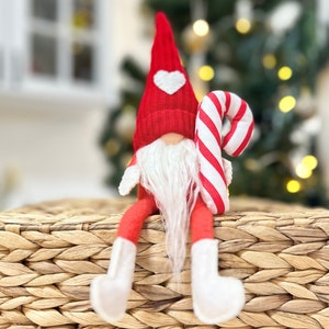 May include: A red and white gnome with a white beard and a candy cane in its hand. The gnome is sitting on a woven basket.