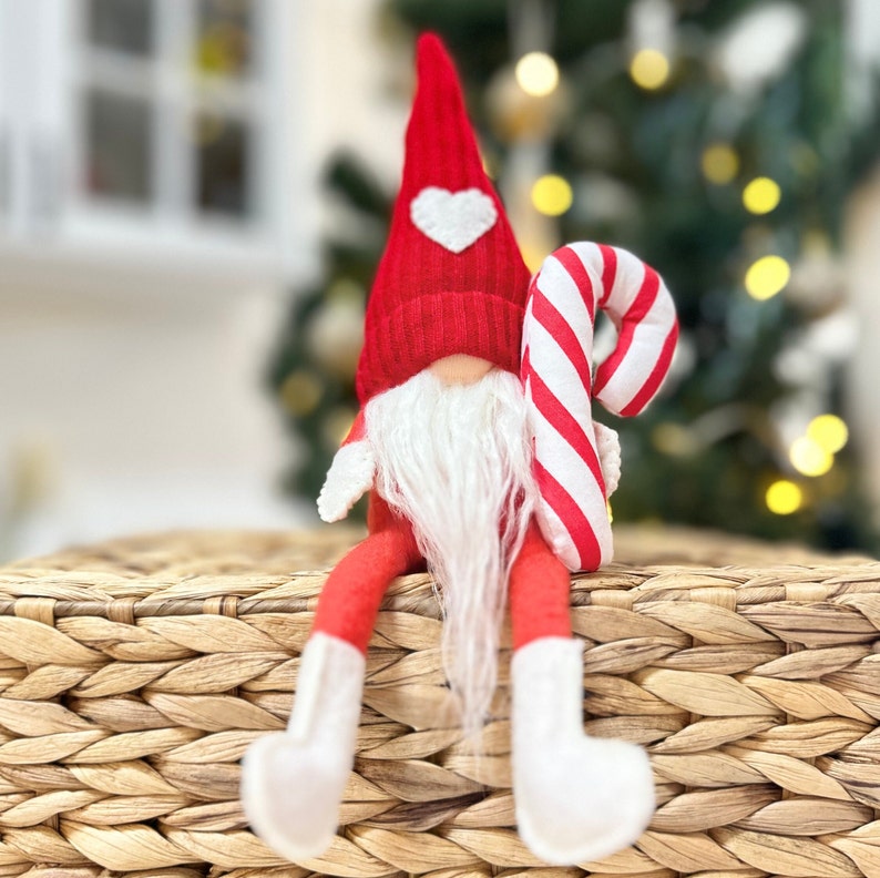 May include: A red and white gnome with a white beard and a candy cane in its hand. The gnome is sitting on a woven basket.