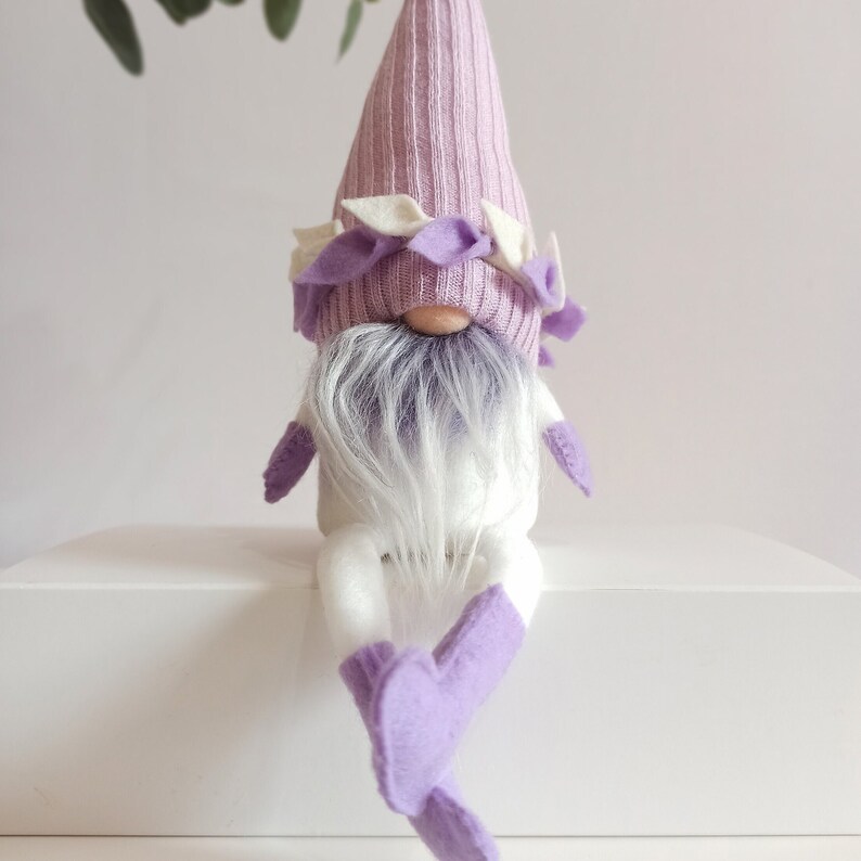 May include: A purple gnome with a white beard and a purple hat with white trim. The gnome is sitting on a white surface.