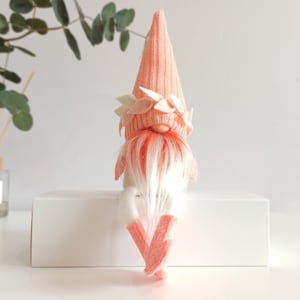 May include: A pink gnome with a white beard and a pink hat with white felt leaves. The gnome is sitting on a white surface.