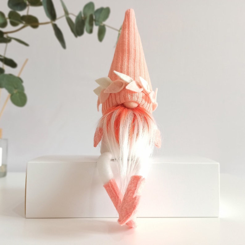 May include: A pink gnome with a white beard and a pink hat with white felt leaves. The gnome is sitting on a white surface.