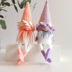 May include: Two gnomes with white beards and white bodies. One gnome is wearing a pink hat with white and pink trim. The other gnome is wearing a purple hat with white and purple trim. Both gnomes are sitting on a white surface.