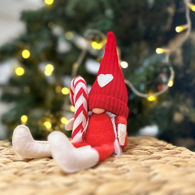 May include: A red felt gnome with a white heart on its hat holds a candy cane. The gnome is sitting on a woven wicker surface.