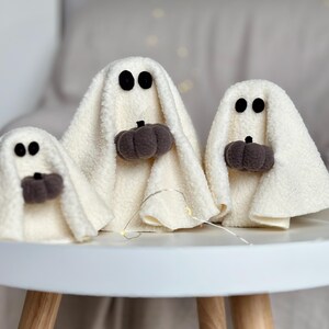 May include: Three white ghost-shaped plush toys with black eyes and holding brown pumpkin-shaped decorations. The ghosts are sitting on a white table with a wooden base.