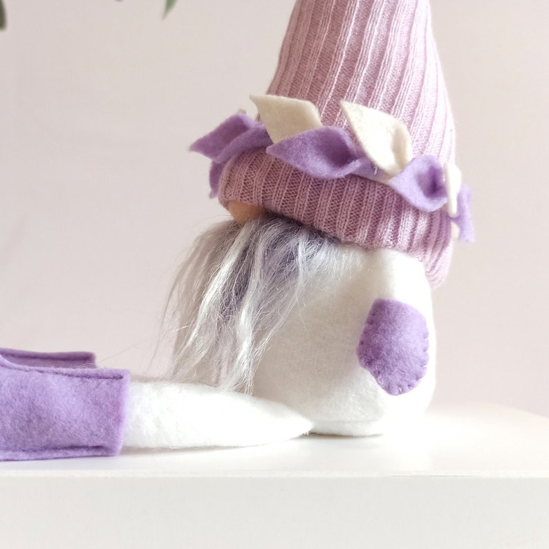 May include: A purple and white gnome with a long white beard and a purple hat with white accents. The gnome is sitting on a white surface.