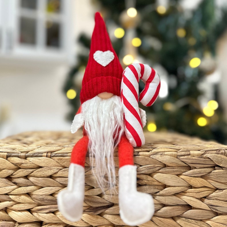 May include: A red and white gnome wearing a red hat with a white heart on it. The gnome is holding a red and white candy cane. The gnome is sitting on a woven basket.