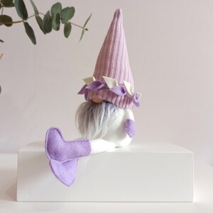 May include: A purple and white gnome with a long white beard and purple felt boots. The gnome is sitting on a white surface.
