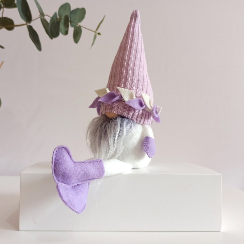 May include: A purple and white gnome with a long white beard and purple felt boots. The gnome is sitting on a white surface.