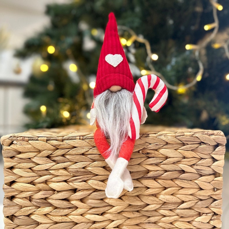 May include: A red and white gnome figurine with a candy cane in its hand. The gnome is wearing a red hat with a white heart on it and has a white beard. The gnome is sitting on a woven basket.