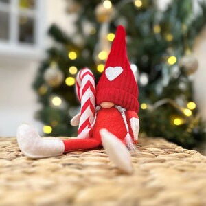 May include: A red and white striped candy cane gnome with a red hat and a white heart on the front. The gnome is sitting on a woven wicker surface.