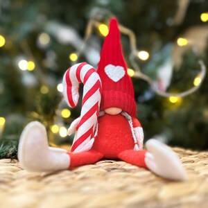 May include: A red and white candy cane gnome wearing a red hat with a white heart. The gnome is sitting on a woven surface.
