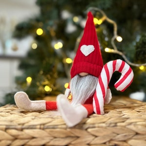 May include: A red and white gnome with a white heart on its hat, holding a red and white candy cane. The gnome is sitting on a woven wicker surface.