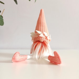 May include: A pink gnome with a white and orange beard, wearing a pink hat with a white flower crown. The gnome is sitting with its legs outstretched.