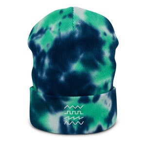 Op de afbeelding: Een tie-dye beanie in tinten van teal, marineblauw en wit. De hoed heeft een omgeslagen rand en een wit geborduurd ontwerp van drie gestapelde golfachtige patronen.