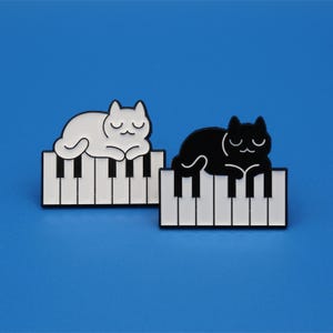 Może przedstawiać: Dwie szpilki emaliowane przedstawiające czarne i białe koty śpiące na klawiaturze fortepianu. Koty są w prostym stylu kreskówkowym.