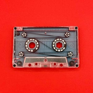Op de afbeelding: Een transparante cassetteband, die de interne componenten laat zien, waaronder twee cirkelvormige spoelen en een magnetische band. De cassette staat tegen een felrode achtergrond, wat een sterk contrast creëert. Het ontwerp van de tape is minimalistisch, met de nadruk op de mechanische elementen.