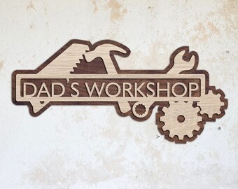 May include: Wooden sign with the text "Dad's Workshop" in a brown wood frame. The sign is decorated with a saw, hammer, wrench, and gears. The sign is 40 cm wide.