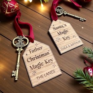Könnte beinhalten: Zwei dekorative Schlüssel mit Holzanhängern und roten Bändern. Die Anhänger tragen die Aufschrift "Santa's Magic Key" und "Father Christmas' Magic Key", mit Namen und Familiennamen. Die Schlüssel sind bronzefarben, und der Hintergrund umfasst Weihnachtsschmuck und Grün.