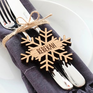 May include: A wooden snowflake place card with the name "REBEKAH" engraved on it. The snowflake is tied to a grey napkin with a fork and knife.