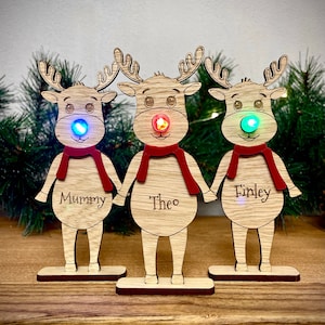 May include: Three wooden reindeer figurines with light-up noses. The reindeer are standing on a wooden surface. The reindeer are decorated with red scarves and have the names "Mummy", "Theo", and "Finley" written on them.