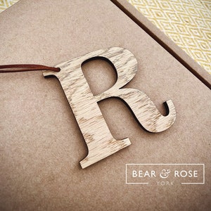 May include: Wooden letter R ornament with a brown leather cord. The letter is made of light brown wood with a natural grain pattern. The ornament is on a brown background with the text "BEAR & ROSE YORK" in white.