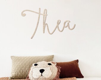 May include: A wooden sign with the name "Thea" written in cursive script. The sign is made of oak wood and is 60 cm wide. The sign is hanging on a white wall with a string of colorful flags above it.