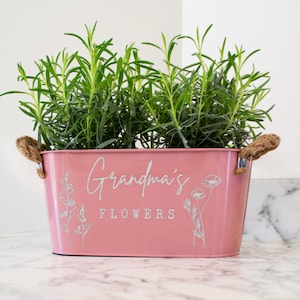 May include: A pink metal planter with rope handles. The planter has the text "Grandma's Flowers" printed on the front in white.