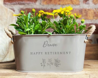 May include: A gray metal planter with rope handles, personalized with the name "Denise" and the text "Happy Retirement". The planter is filled with yellow flowers and green foliage.