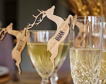 Puede incluir: Porta tarjetas de lugar de madera en forma de ciervo con nombres escritos en ellos. Las tarjetas de lugar están sujetas a copas de vino.