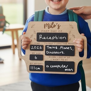 May include: A child smiles, holding a wooden sign that reads "Milo's First Day of Reception." The sign displays the year 2026, age 4, height 111cm, and interests: trains, planes, and dinosaurs. The child's ambition is to be a computer man.