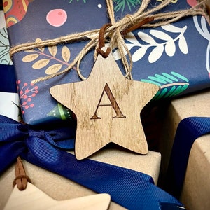 May include: A close-up of a wooden star ornament with the letter "A" engraved on it. The star is tied with twine to a blue and white patterned gift box.