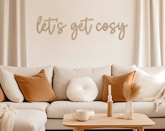 May include: A white sectional sofa with brown throw pillows and a white throw blanket. A wooden coffee table with a white mug and a candle holder. The wall behind the sofa has a wooden sign that reads "Let's get cosy."