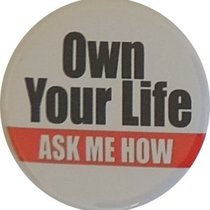 May include: White button with black text that reads "Own Your Life ASK ME HOW" with a red stripe at the bottom.