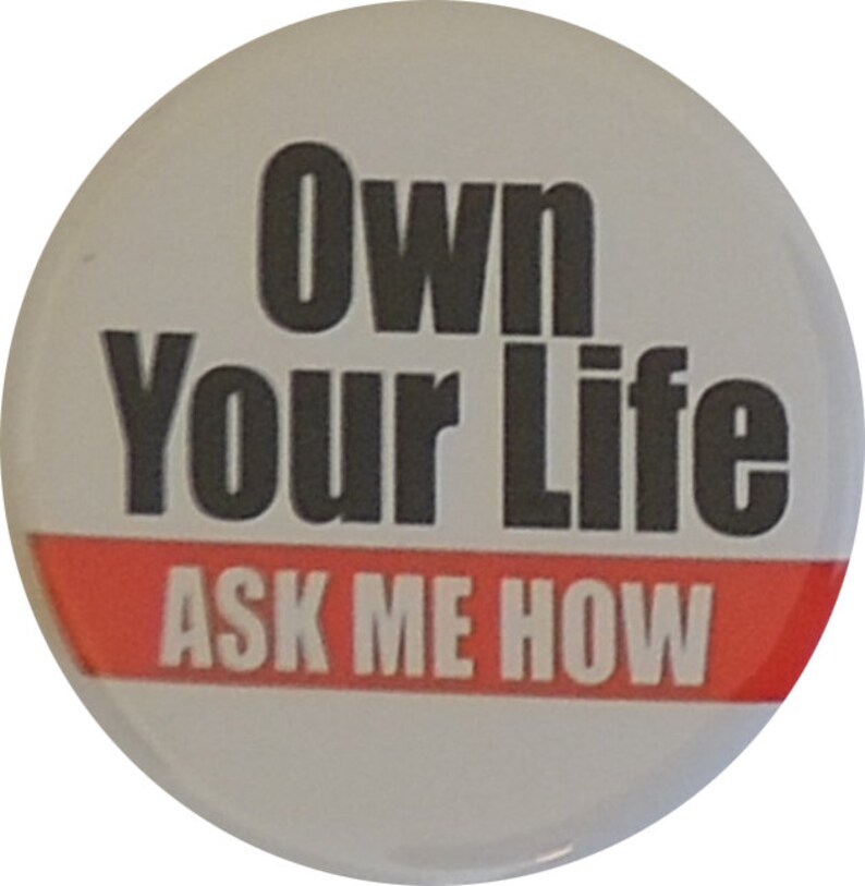 May include: White button with black text that reads "Own Your Life ASK ME HOW" with a red stripe at the bottom.