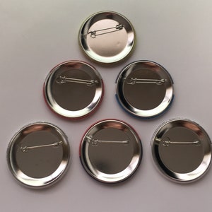 May include: Six oval-shaped metal button badges with different coloured borders. The badges are silver with a safety pin fastening on the back.