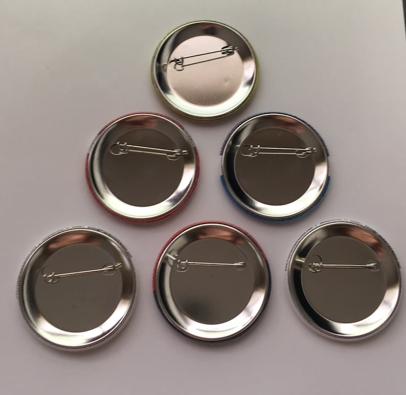 May include: Six oval-shaped metal button badges with different coloured borders. The badges are silver with a safety pin fastening on the back.