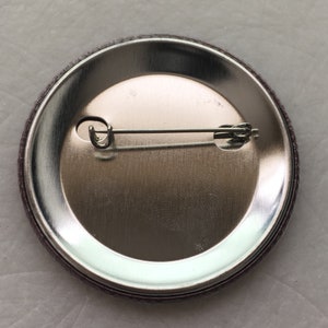 May include: The back of a round button with a silver metal backing and a pin attached.